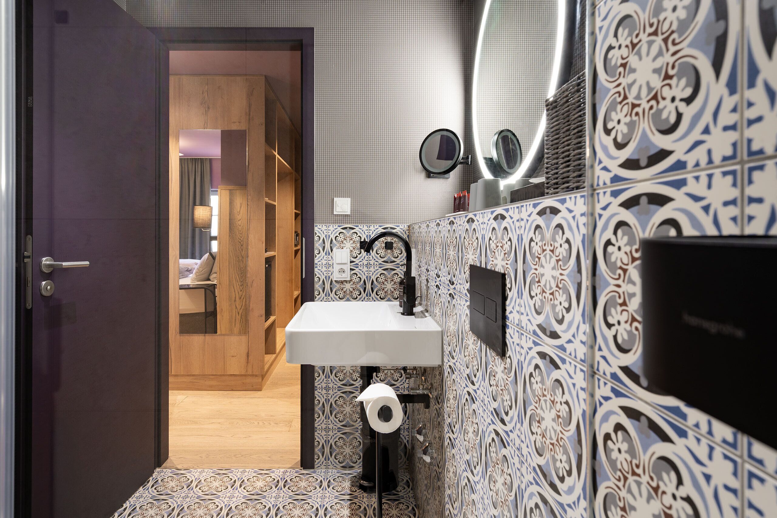 Een badkamer met een witte wastafel, betegelde vloer, een spiegel en een toiletrolhouder.