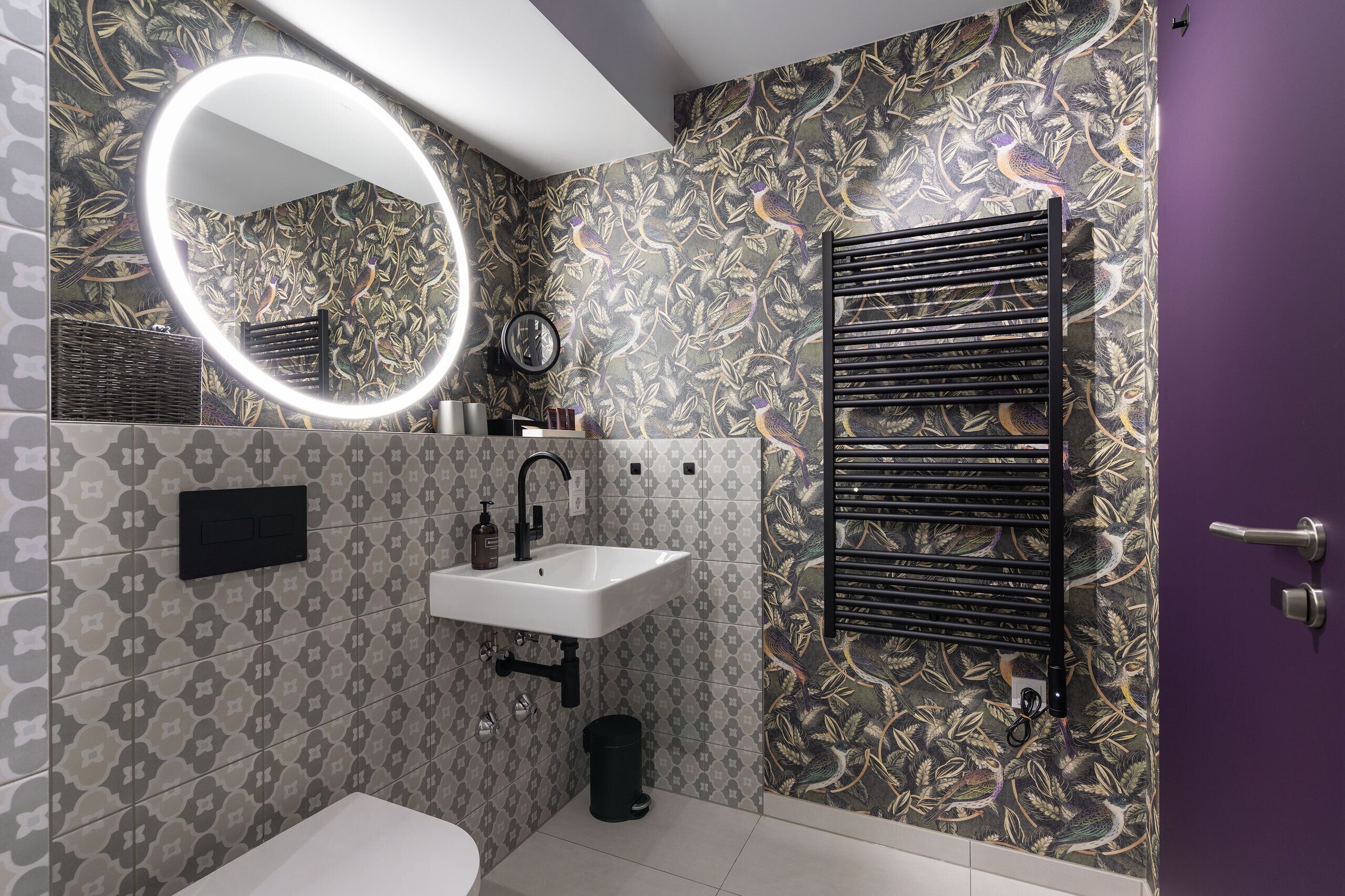 Un baño con un lavamanos blanco, un espejo con luz, un perchero para toallas y un papel tapiz estampado.