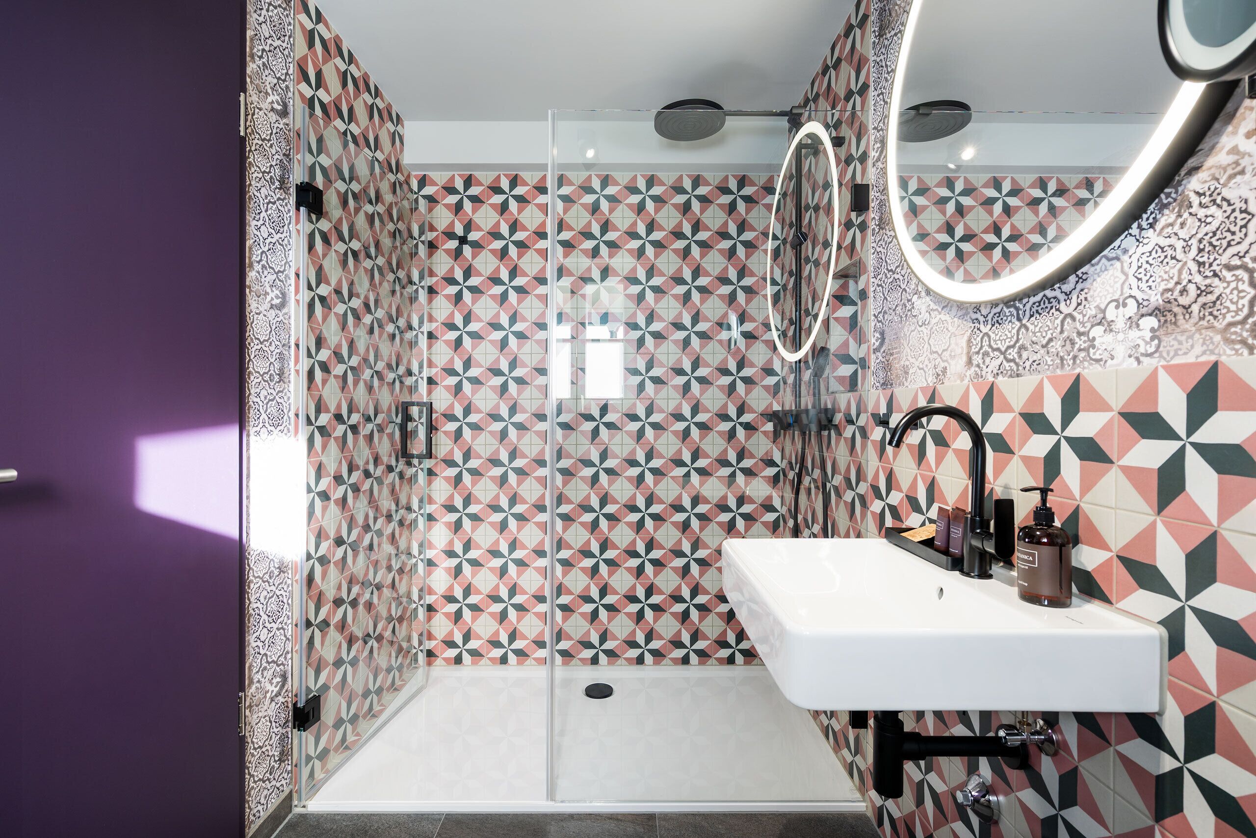 Une salle de bain avec un mur recouvert de carreaux aux motifs géométriques, un lavabo blanc avec un miroir au-dessus, et un espace douche.