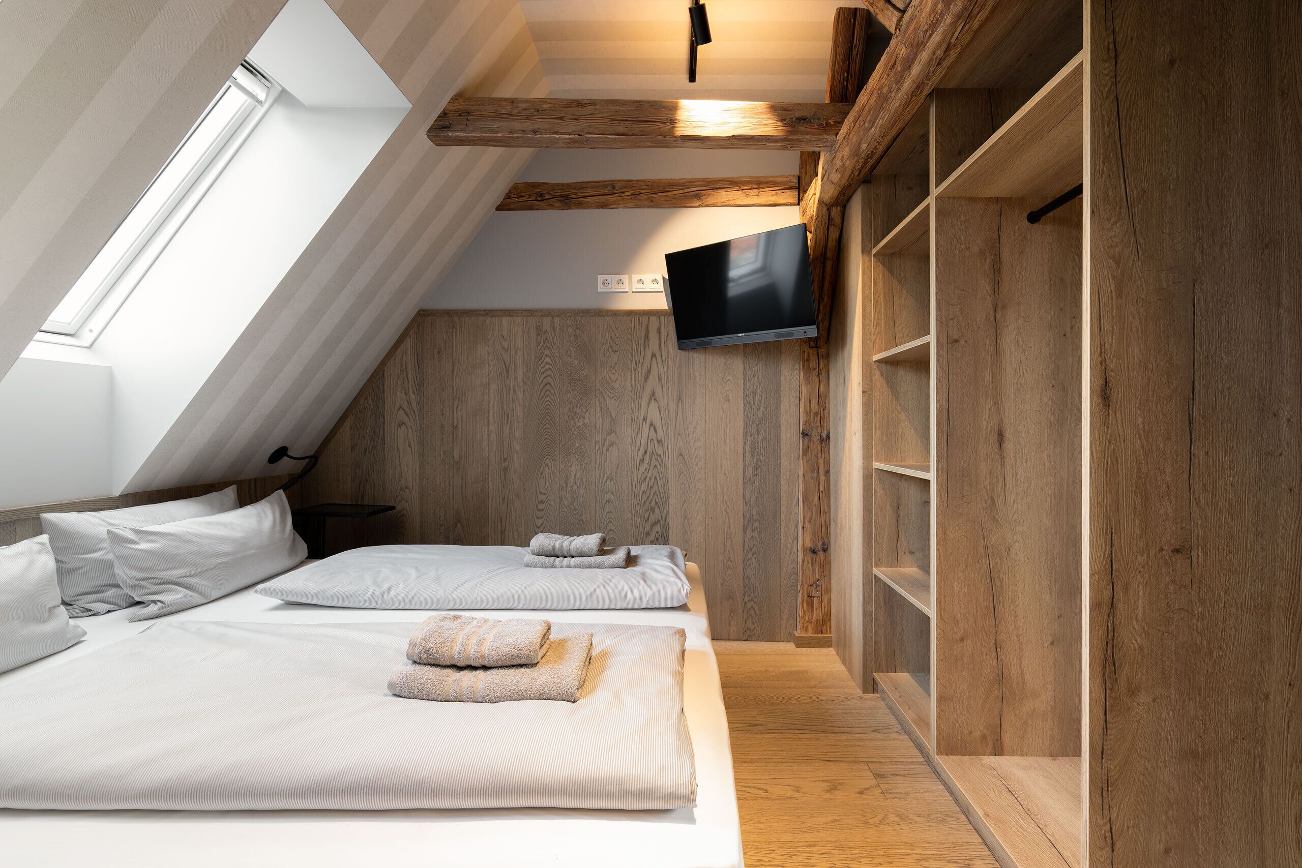 A bedroom with a sloped ceiling, wooden beams, a flat-screen TV, and a bed with neatly folded towels.