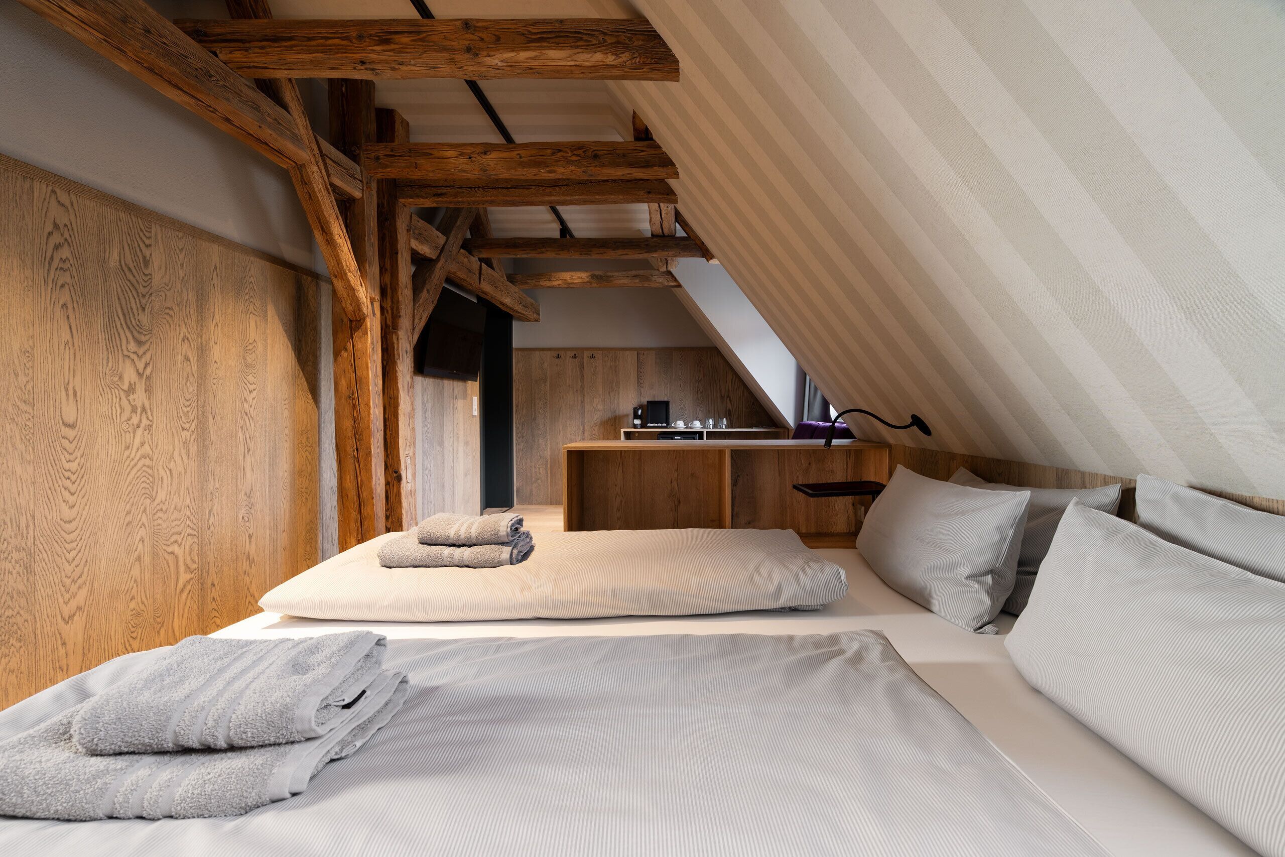 A bedroom with a sloped ceiling, wooden beams, a bed with pillows and towels, and a kitchenette in the background.