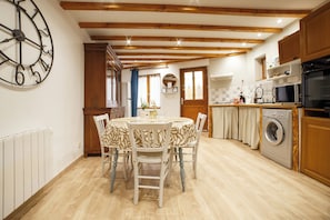 A kitchen with wooden beams, a dining table with chairs, a washing machine, and a clock on the wall. - Charming stone house with terrace & barbecue, near valence (Montélier)