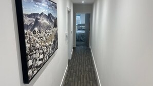 A hallway with a large framed picture of a mountainous landscape on the left, a door at the end, and a bed visible in the background. - Chapel Rise Apartments (Long Eaton)