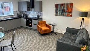 A modern kitchen with grey cabinets, a black range hood, and a window. There is a grey sofa, an orange armchair, and a small table with a lamp. - Chapel Rise Apartments (Long Eaton)