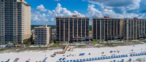 Un hotel frente a la playa con varios edificios altos, una playa arenosa con numerosas sombrillas y un cielo azul despejado.