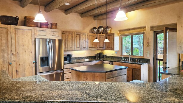 Cocina con encimeras de granito, armarios de madera, refrigerador de acero inoxidable e iluminación colgante.