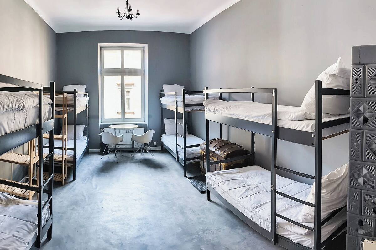 A dormitory room with bunk beds, a small table, and chairs, a window with natural light, and a chandelier.