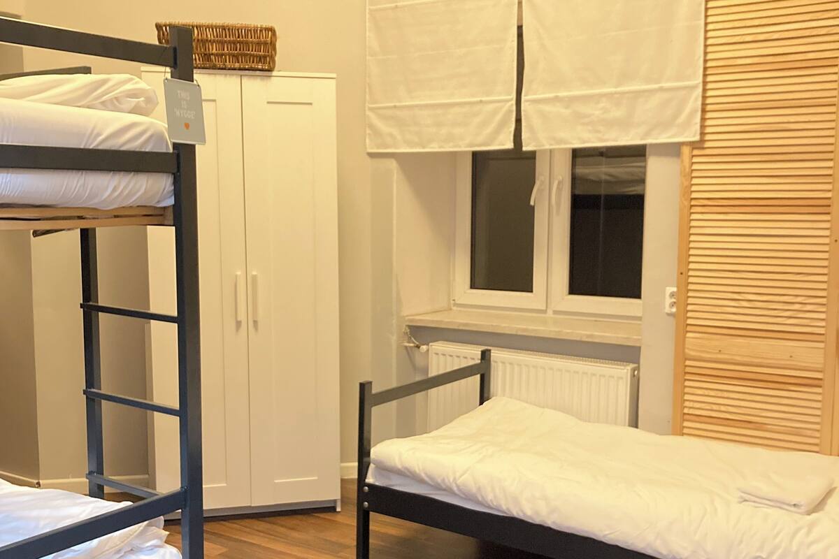 A dormitory room with bunk beds, a wardrobe, a window with blinds, and a radiator.