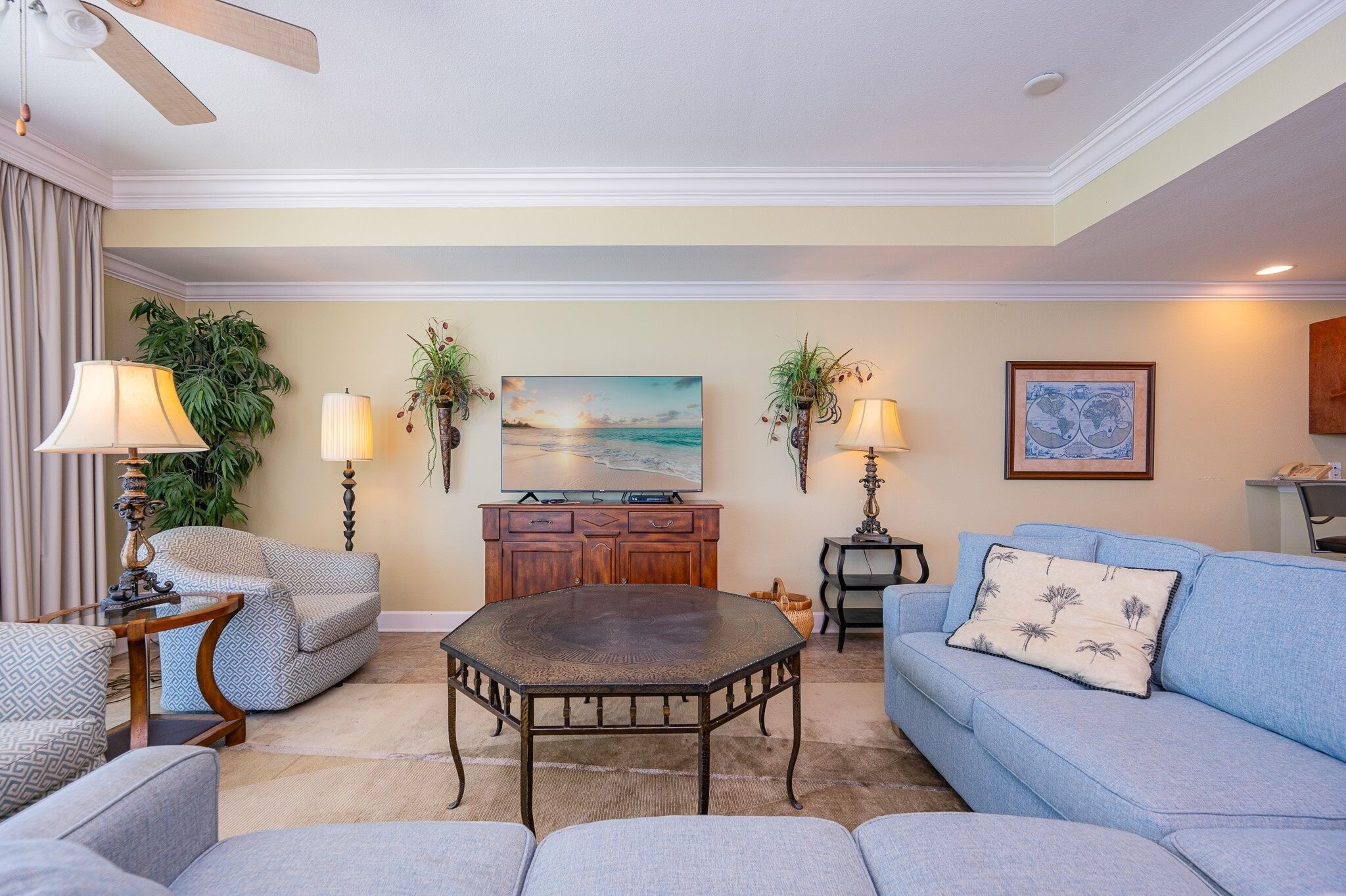 A living room with a sofa, armchairs, a coffee table, a TV, decorative plants, and a ceiling fan.