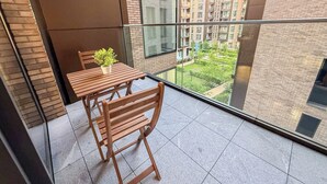 Un balcón con mesa y sillas de madera, una planta en maceta y vistas a una zona residencial con edificios y zonas verdes.