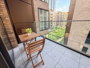 Un balcone con tavolo e sedie in legno, una pianta in vaso e vista su una zona residenziale con edifici e vegetazione.