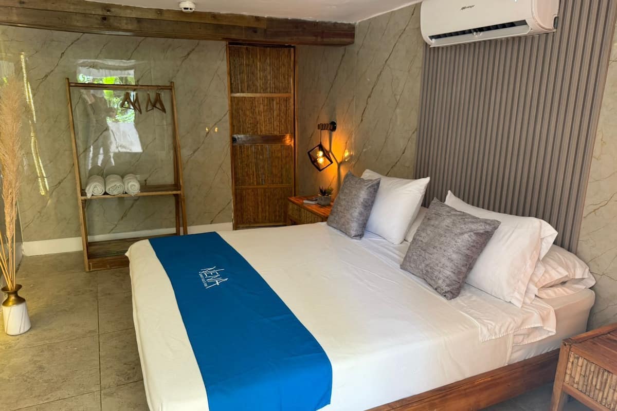 A hotel room with a large bed, a wooden headboard, a wall-mounted air conditioner, a shelf with towels, and a mirror with a wooden frame.