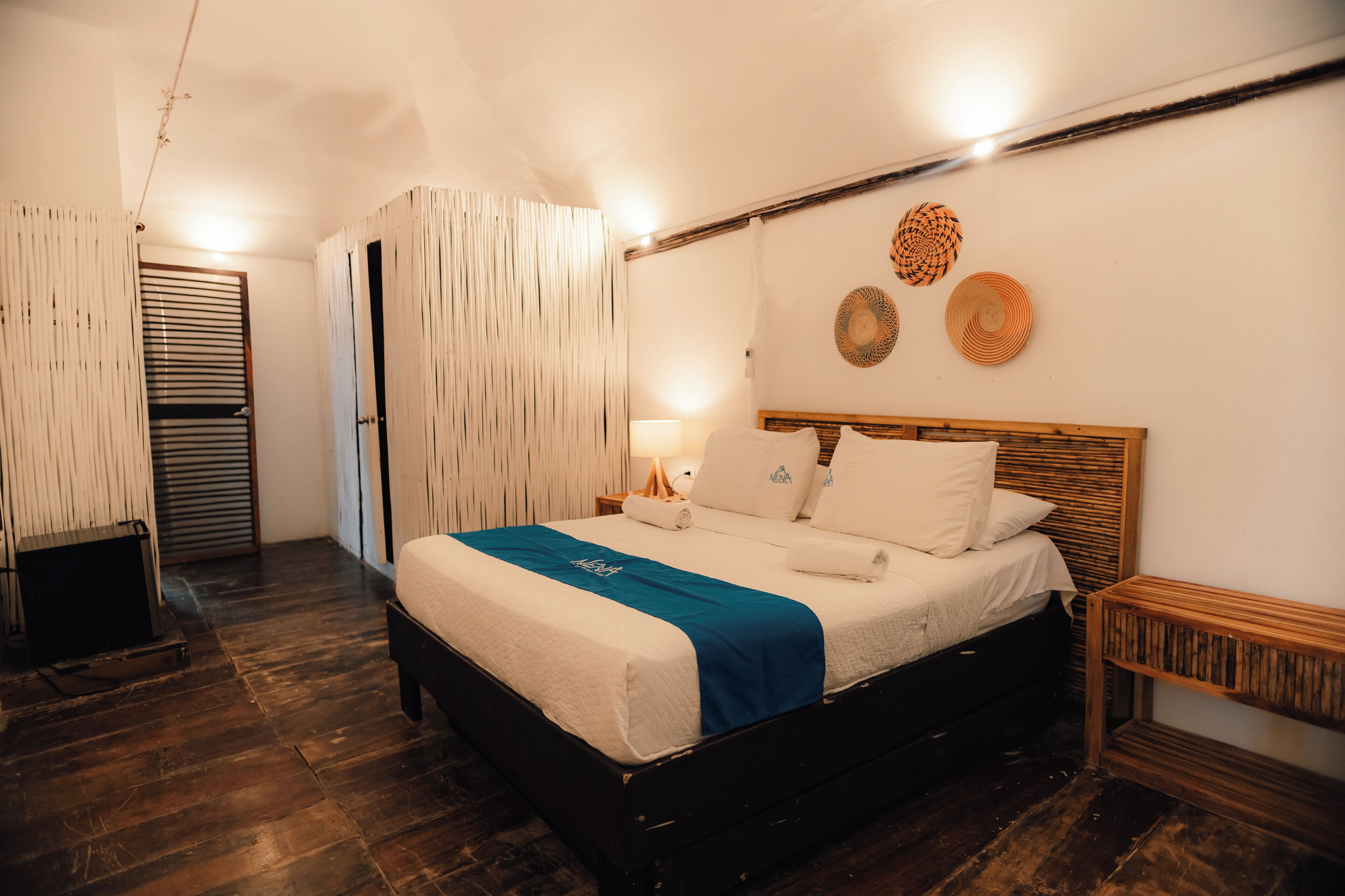 Un dormitorio con cama de madera, ropa de cama blanca, una manta azul, una mesita de noche de madera y una lámpara de pared.