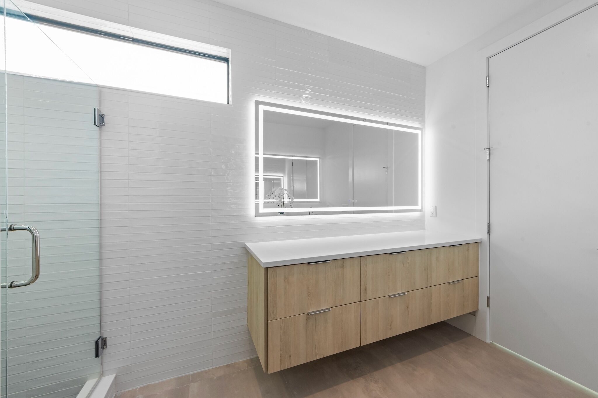 Un baño moderno con cabina de ducha de vidrio, tocador de madera con espejo y pared de azulejos blancos.