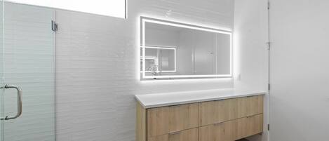 Un baño moderno con cabina de ducha de vidrio, tocador de madera con espejo y pared de azulejos blancos.