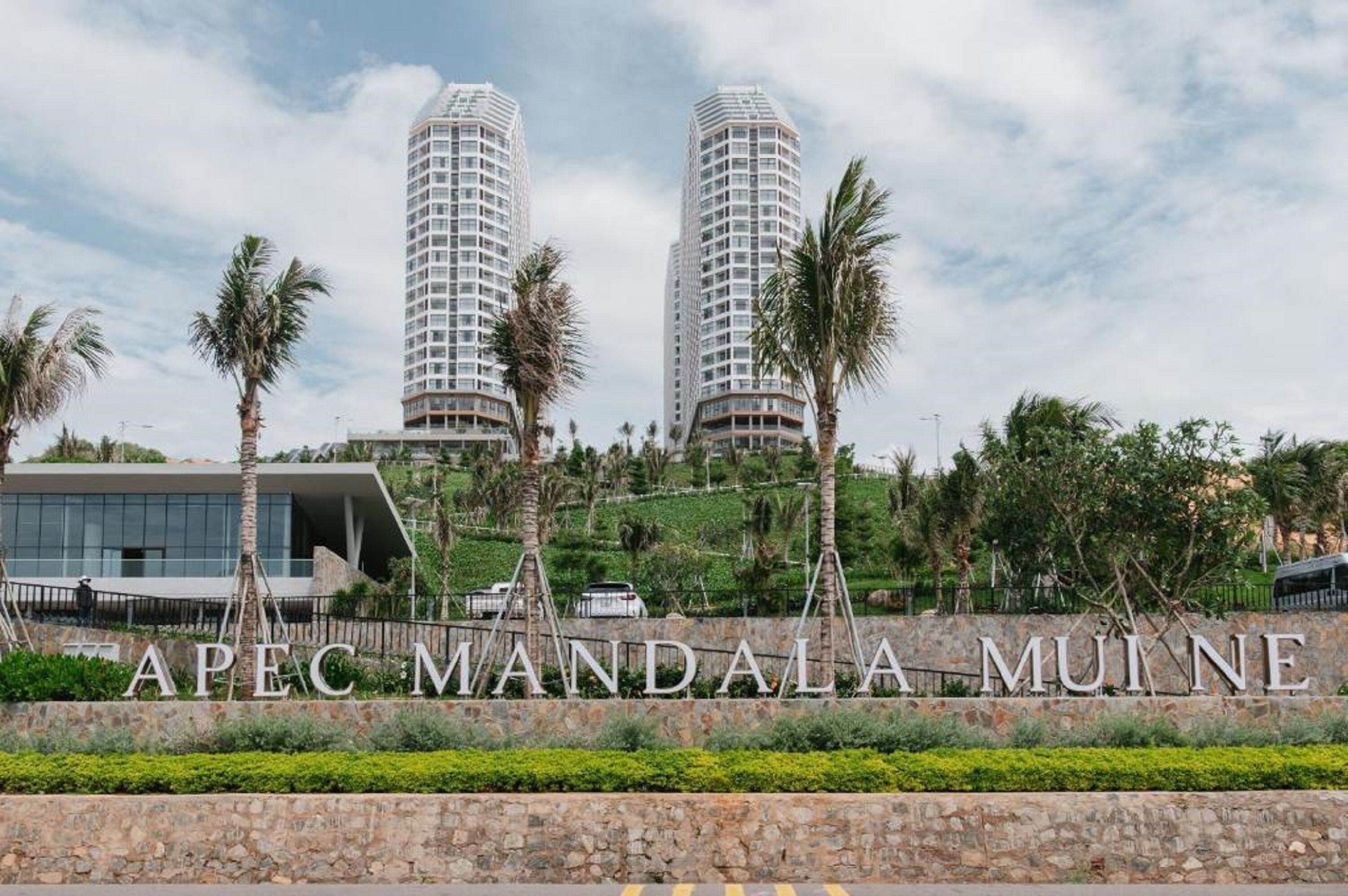 A modern building complex with two tall towers, a landscaped area with palm trees, and a sign reading "APEC MANDALA MUINE".