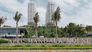 A modern building complex with two tall towers, a landscaped area with palm trees, and a sign reading "APEC MANDALA MUINE".