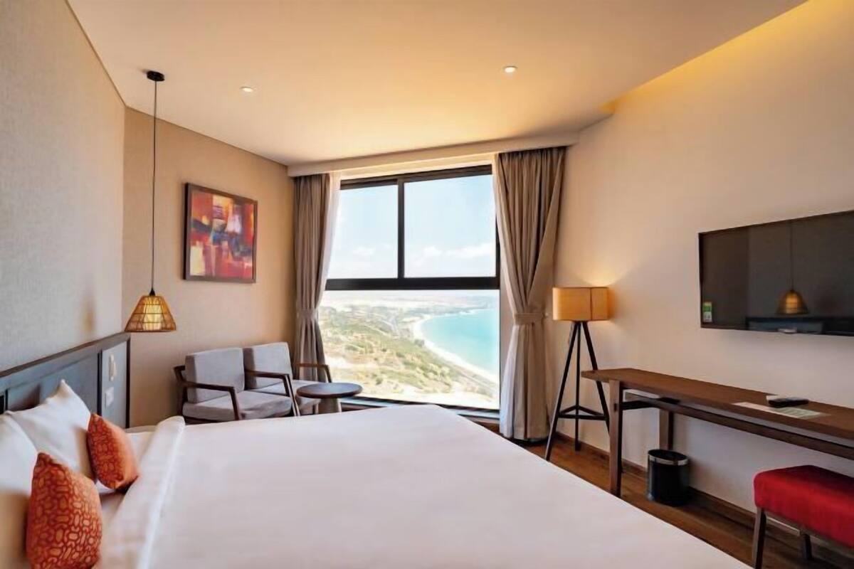 Une chambre d’hôtel équipée d’un lit, d’un bureau, d’une chaise, d’une petite table, d’une lampe, d’une télévision et d’une grande fenêtre donnant sur la plage.