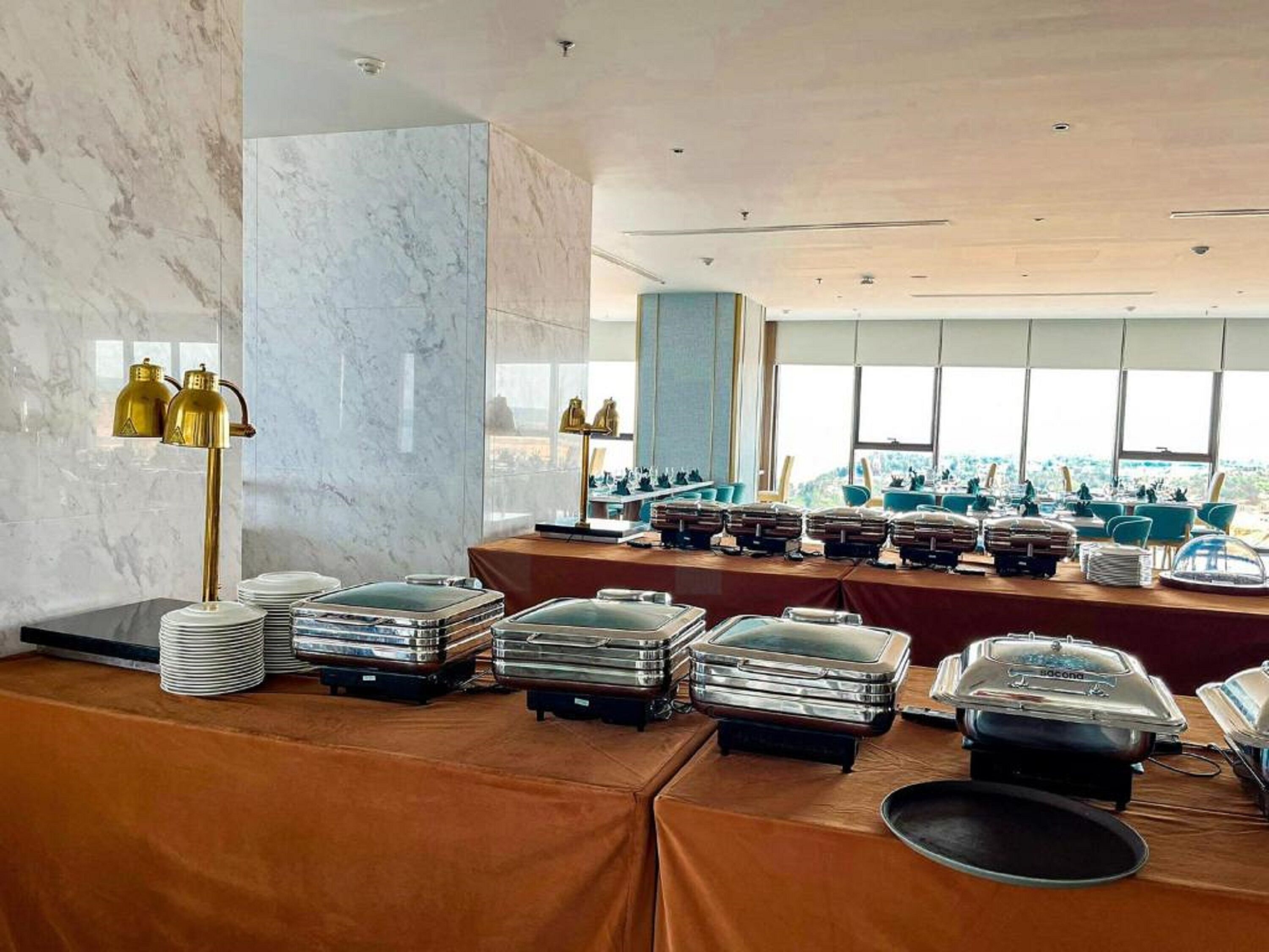 A buffet setup with multiple serving stations, a marble wall, and a view of the city through large windows.