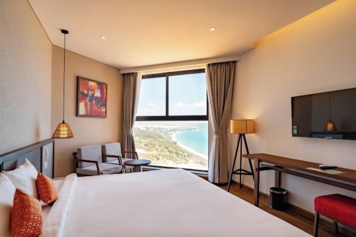 Hotelkamer met een bed, bureau, stoel, een klein tafeltje, lamp, televisie en een groot raam met uitzicht op het strand.