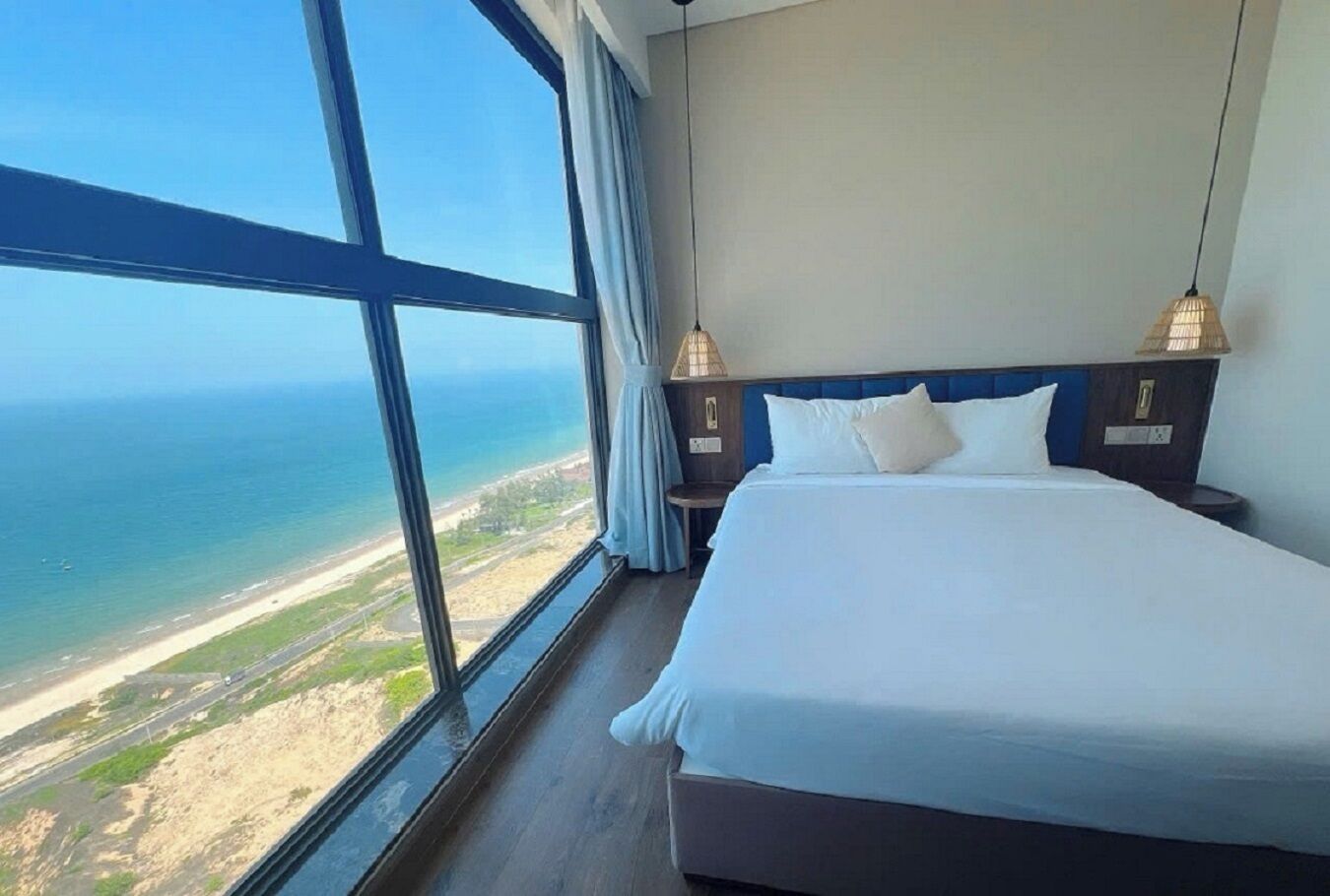 A hotel room with a large window offering a view of the ocean and a road. The room has a bed with white bedding and two bedside lamps.