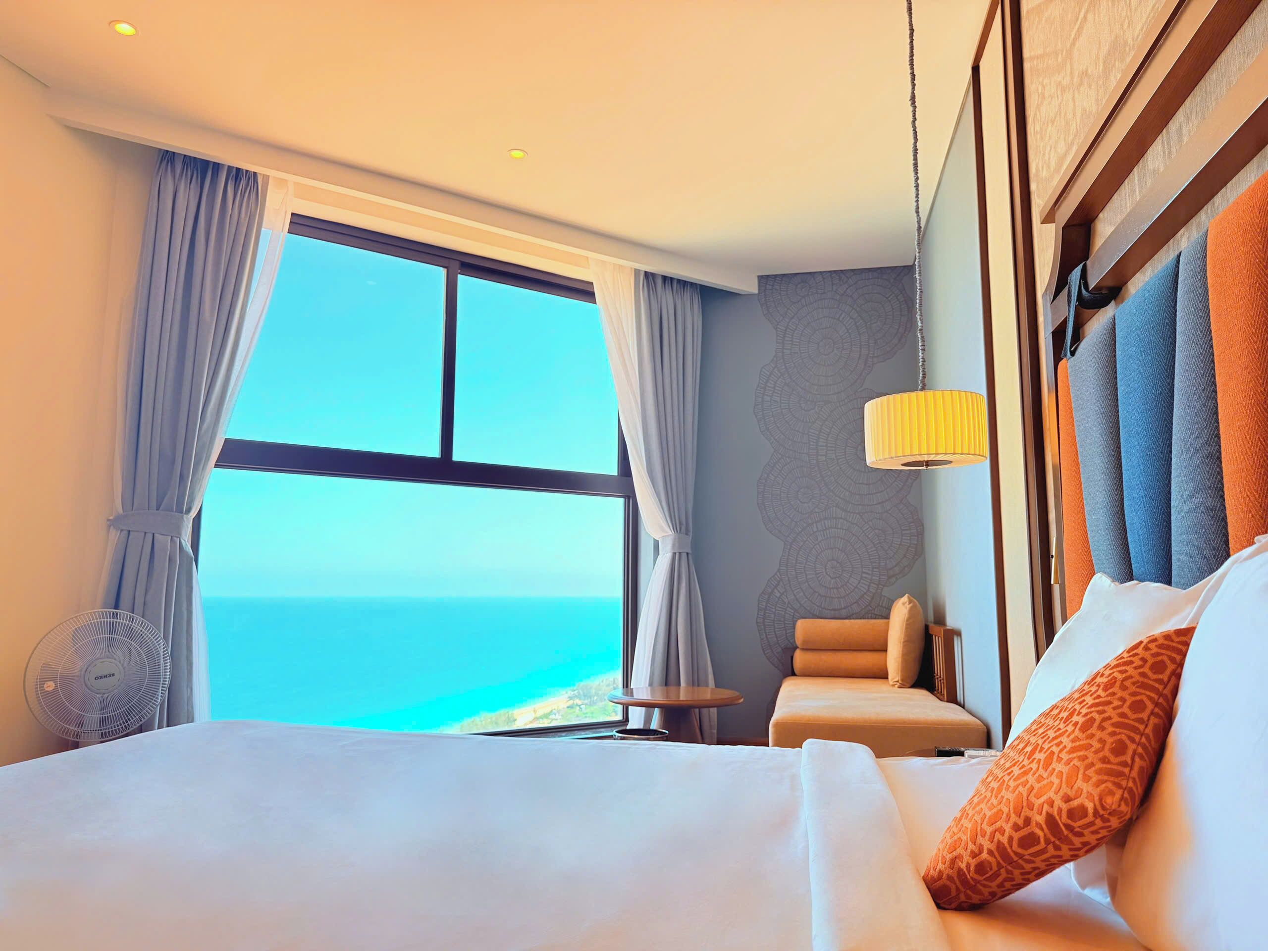 Habitación de hotel con una cama grande, una silla, una mesita, un ventilador y vista al océano.