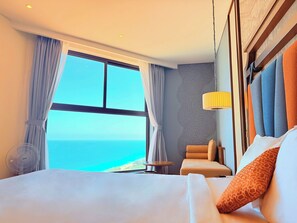 Une chambre d’hôtel avec un grand lit, une chaise, une petite table, un ventilateur et une vue sur l’océan.