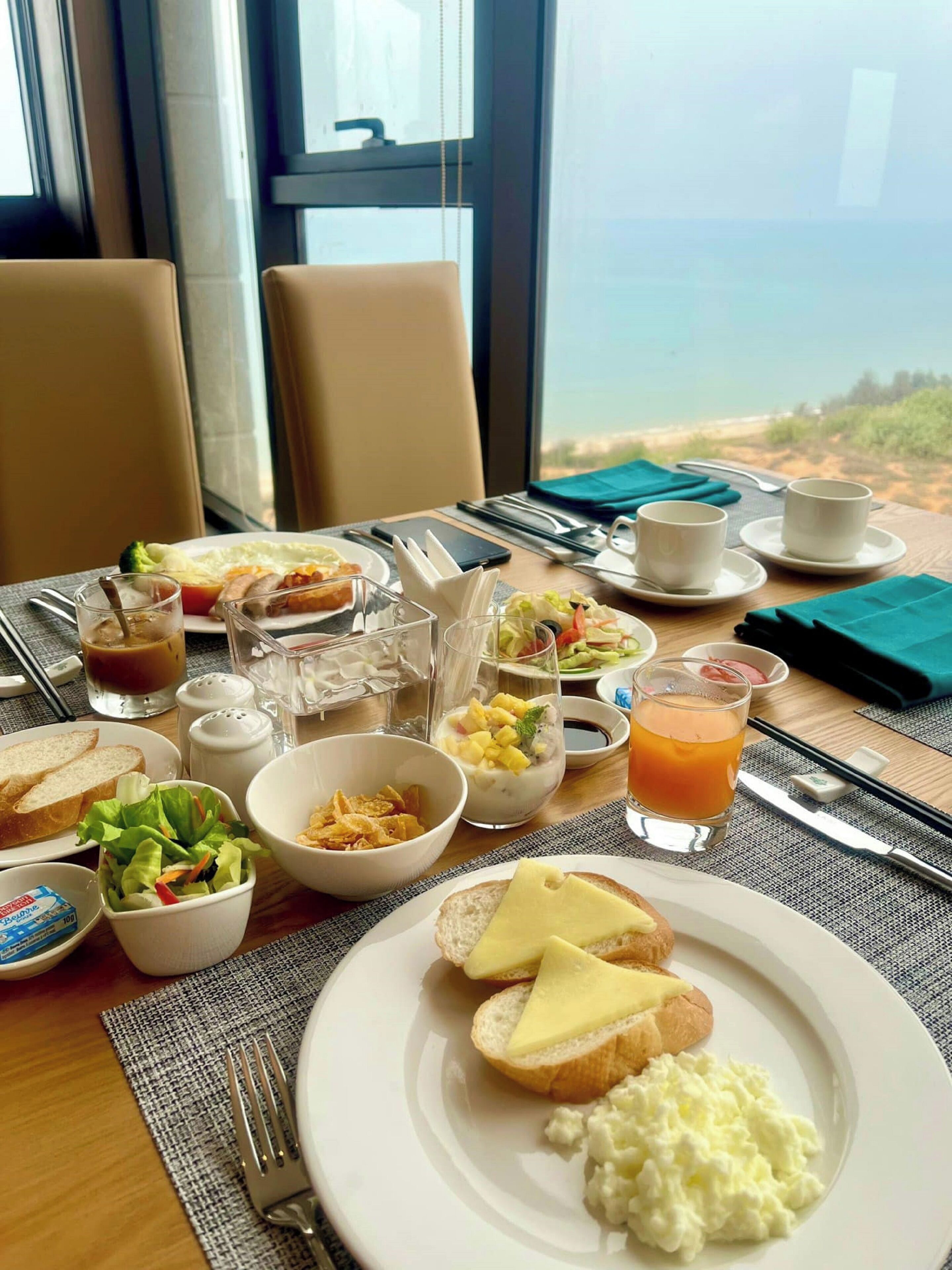 A breakfast setting with various dishes, including toast, scrambled eggs, and a salad, on a table with a view of the beach.