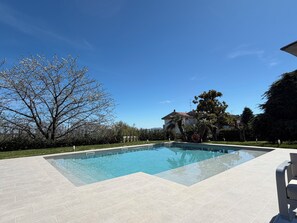 A swimming pool with a tiled deck, surrounded by trees and a house in the background.