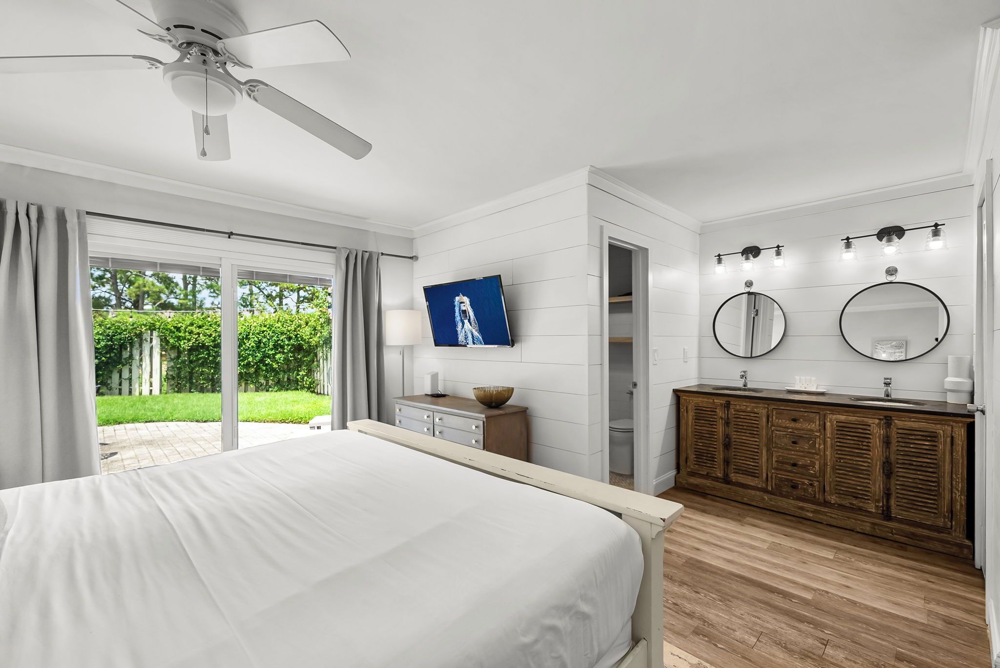 Een slaapkamer met een bed, een plafondventilator, een televisie, een houten dressoir en een badkamer met twee wasbakken en spiegels.