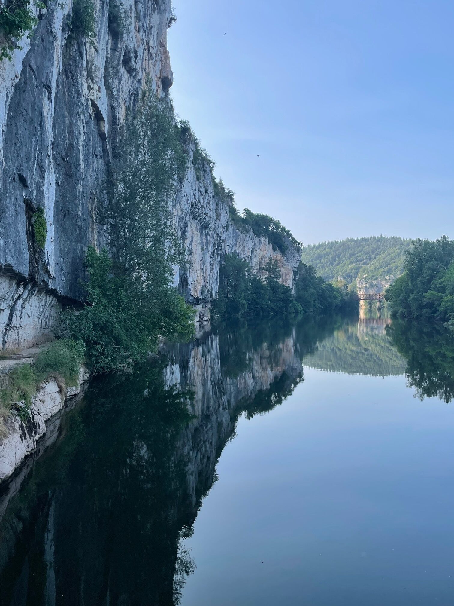 A calm river reflecting a rocky cliff and a distant bridge.