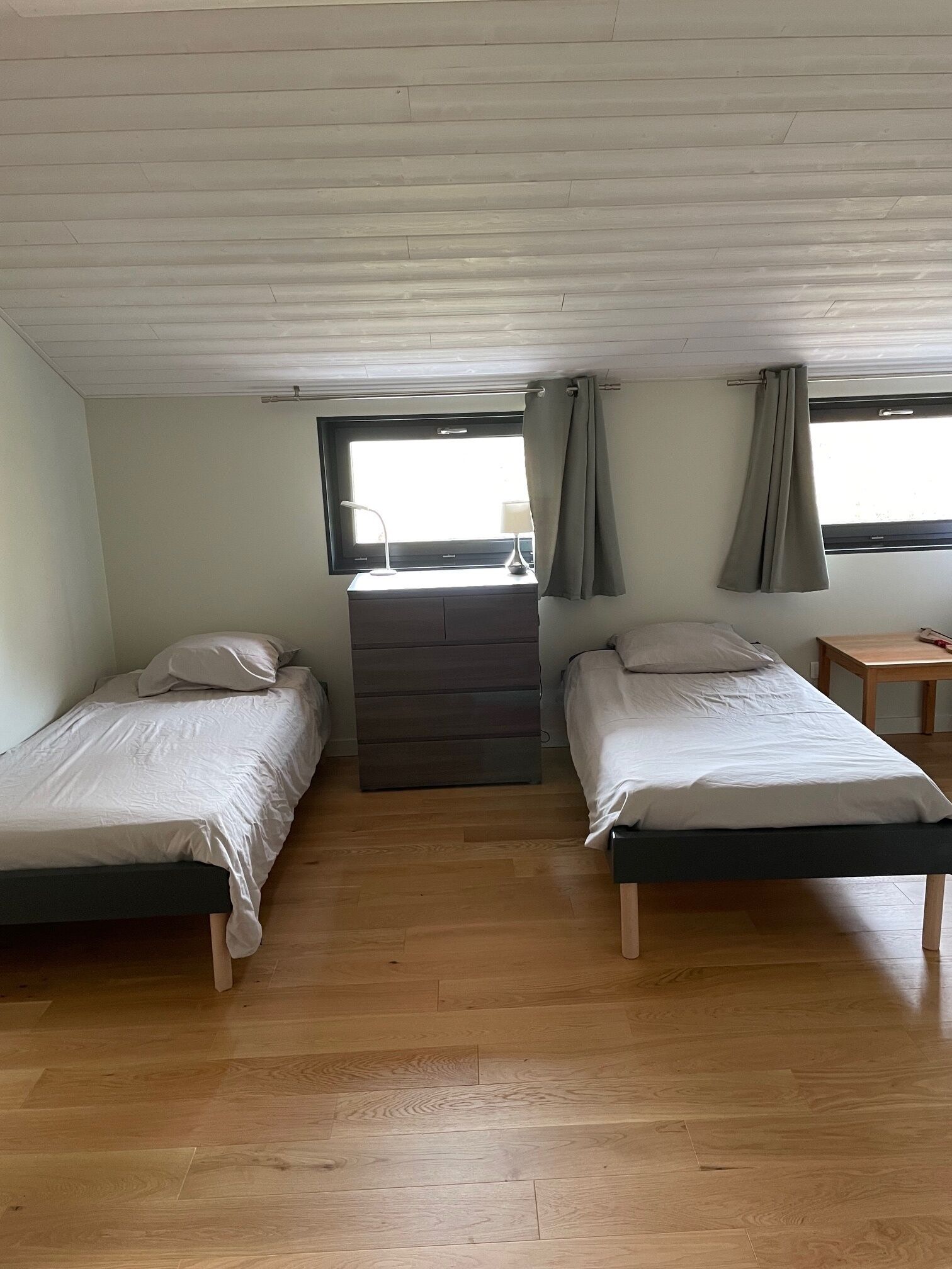 Een kamer met twee eenpersoonsbedden, een dressoir, een raam met gordijnen en een klein tafeltje.