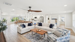 A spacious living area with a large sofa, a coffee table, and a patterned armchair. There is a dining area in the background with a table and chairs.