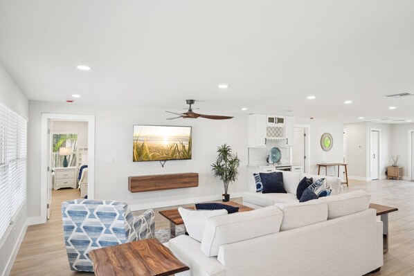 A spacious living room with a white sofa, a patterned ottoman, and a wooden coffee table. A ceiling fan and recessed lighting are visible.