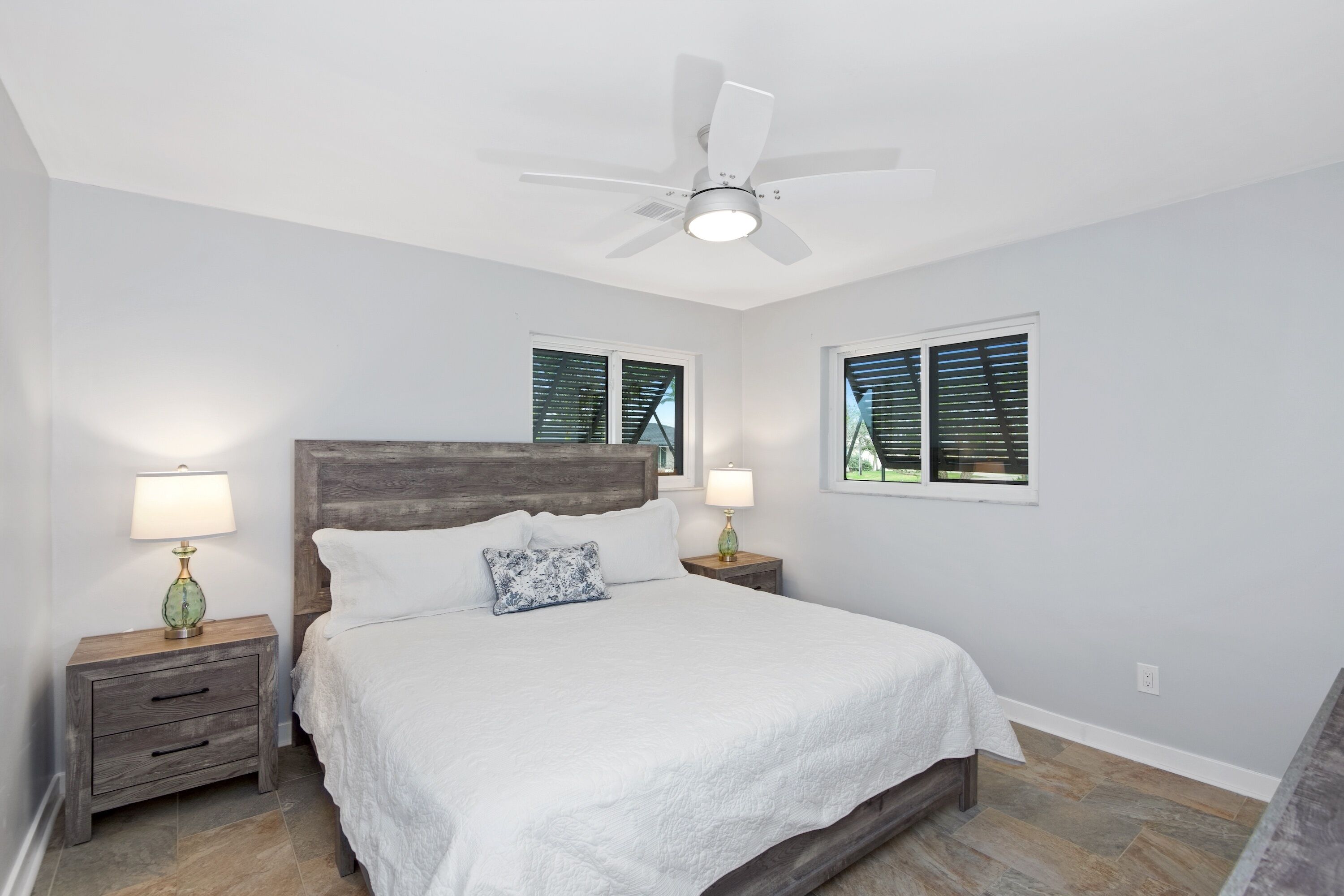 A bedroom with a bed, two nightstands, a ceiling fan, and two windows with blinds.