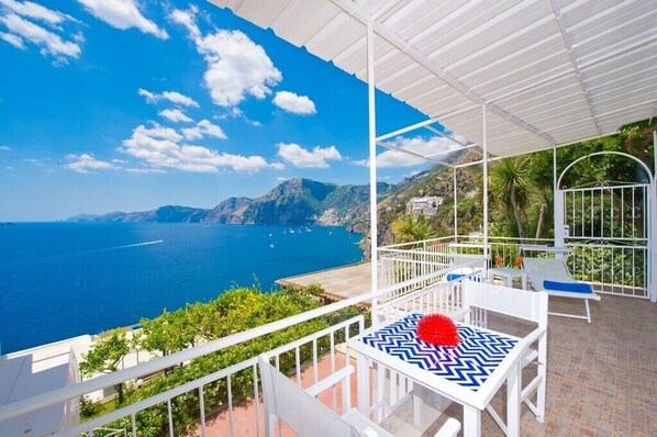 A balcony with a view of the sea and mountains, white furniture, and a patterned tablecloth. - Il Corallo Praiano (Praiano)