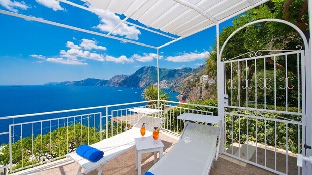 A balcony with a white metal railing, a white table, and two lounge chairs with blue cushions. - Il Corallo Praiano (Praiano)
