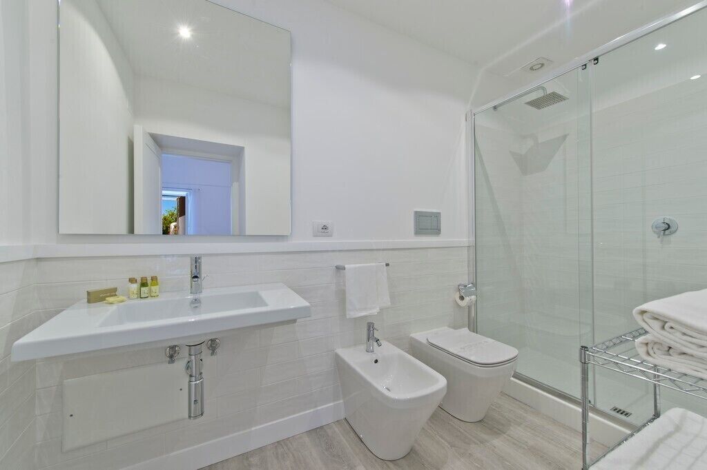 A modern bathroom with a large mirror, a sink, a bidet, a toilet, and a glass-enclosed shower area.