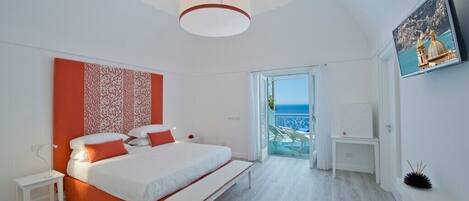 A modern hotel room with a large bed, a bench, a bedside table, a small table, and a view of the sea through a glass door.