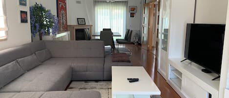 A modern living room with a grey sectional sofa, a glass coffee table, a flat-screen TV on a white stand, and a dining area in the background.
