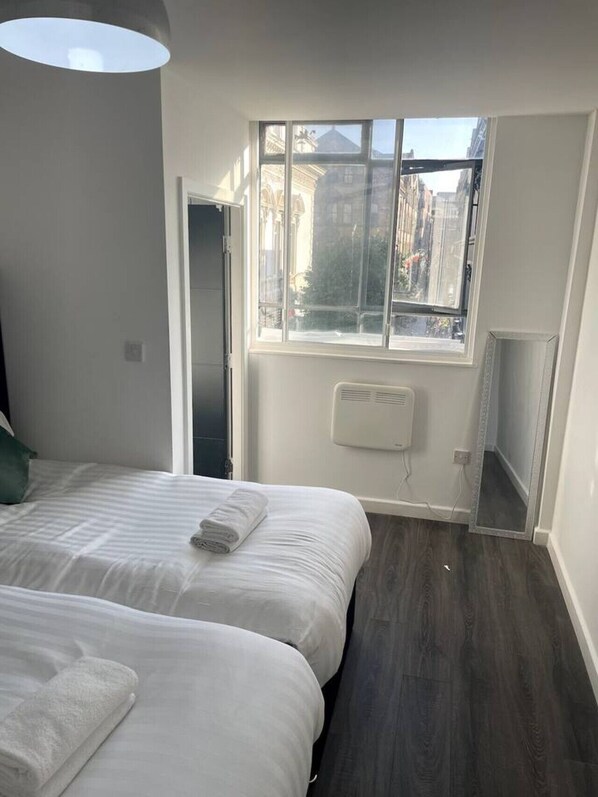 A modern bedroom with two beds, a large window with a view of buildings, and a ceiling light fixture.
