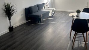 Moderni olohuone, jossa on tumma puulattia, harmaa sohva, pieni pyöreä pöytä, jossa on ruukkukasvi, ja suuri ikkuna, josta tulee runsaasti luonnonvaloa.