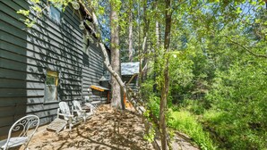 Ein rustikales Blockhaus mit Holzwänden, einer überdachten Sitzgelegenheit mit Metallstühlen und einer naturnahen, bewaldeten Umgebung.