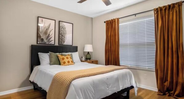 Un dormitorio con cama, mesita de noche, ventilador de techo y ventana con cortinas.