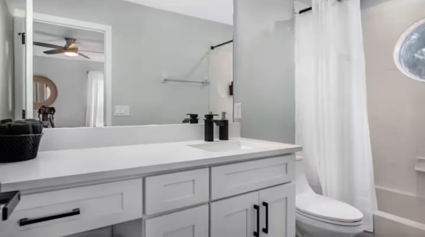 A bathroom with a white vanity, a mirror, a ceiling fan, a toilet, and a shower curtain.