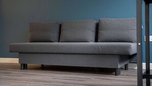 A grey sofa with three cushions against a dark blue wall, placed on a wooden floor.