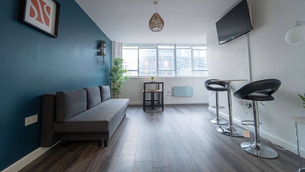 A modern living room with a grey sofa, a small table with a plant, two bar stools, a TV, and a large window with a city view.