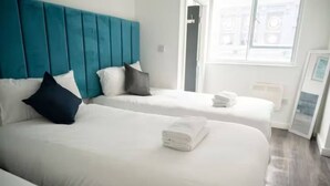 A hotel room with two beds, a teal headboard, white bedding, and a view of the city through the window.