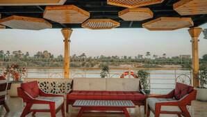 A covered outdoor seating area with wooden furniture and a view of a river and palm trees.