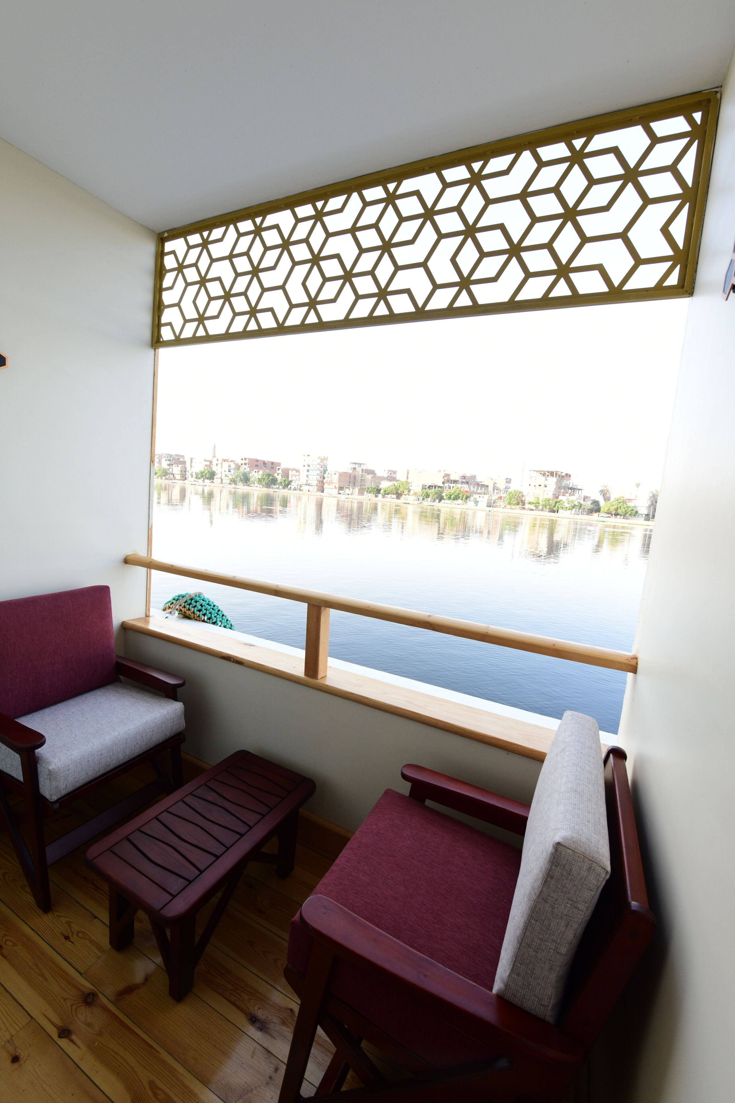 A balcony with two wooden chairs and a bench, overlooking a calm body of water with buildings in the background.